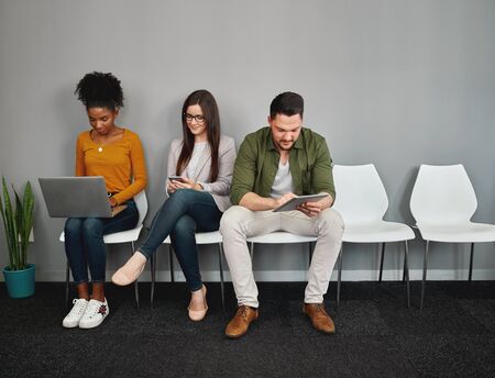 Young multiethnic people sitting on chair chatting and browsing online in smartphones, digital tablet and laptop preparing for interviewの写真素材