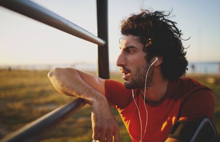Side view of an exhausted young male athlete listening to music on earphones leaning on bars in gym park looking away into the sunsetの写真素材