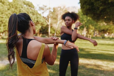 Two sporty fitness young diverse female friends stretching their muscles in park on a summers day - two friends working out togetherの写真素材