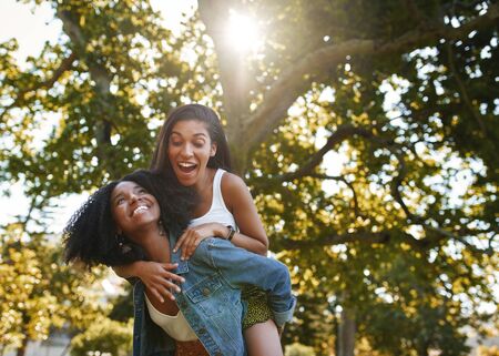 Portrait of a excited young woman enjoying the piggyback ride on her african american friends back in the park having fun and laughingの写真素材