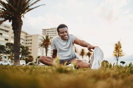 Smiling portrait of a athlete young man sitting on green grass doing stretching exercise at parkの写真素材