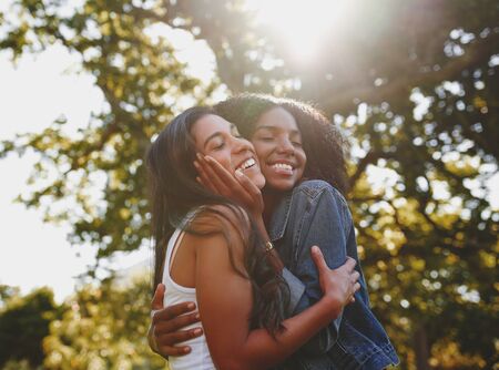 Smiling portrait of a diverse young female with eyes closed cuddling and loving each other in the park with sunlight shining throughの写真素材