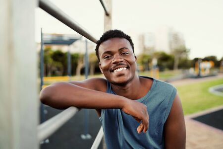 Smiling portrait of a sporty young african american man leaning on bar at the public park looking to...