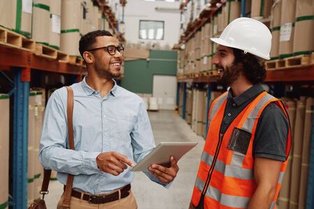 Warehouse manager wearing in formal clothing talking to loader in aisle between tall shelves with packed goods in modern factoryの写真素材