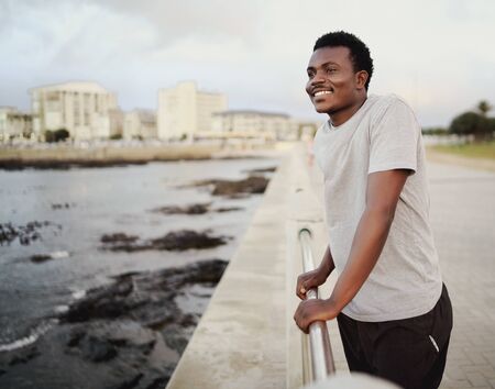 Portrait of a smiling young athlete standing next to a railing looking out at sea relaxing after the workoutの写真素材