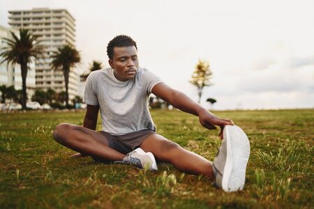 An african american young fitness man sitting on green grass stretching his legs before workoutの写真素材