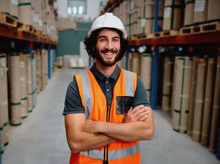 Portrait of cheerful supervisor in a warehouse for delivering and transporting industrial goods wearing white helmet and orange vest uniform standing with folded arms in aisleの写真素材