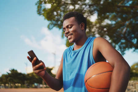 Portrait of a smiling young male basketball player holding ball in hand texting messages on his smart phone at parkの写真素材