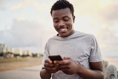 Portrait of a happy athlete young man texting messages on smart phone standing against the city...
