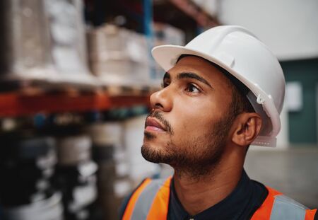 Side close-up of thoughtful african american male worker looking at copy space in warehouse wearing a white helmetの写真素材