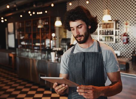 Young cafe owner checking for online order on digital tablet while standing in coffee house wearing...