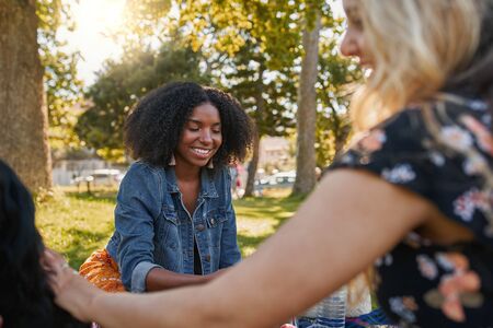 Smiling portrait of a happy african american young woman sitting with her friends in the park having a picnic on a warm summer dayの写真素材
