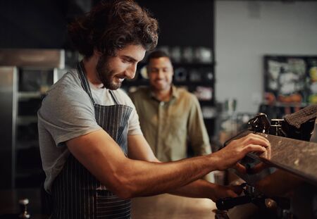Handsome successful male worker making fresh coffee using machine while customer watching through...