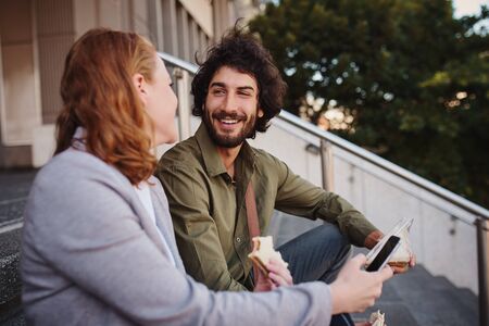 Portrait of young handsome man in conversation with woman during break while eating sandwich for...