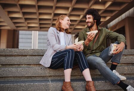 Smiling young man and beautiful woman eating sandwich while in a conversation sitting on stairs