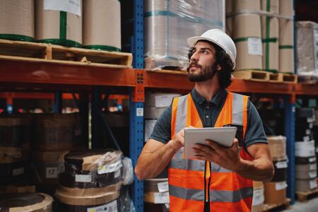 Handsome young bearded man wearing safety vest and hard hat using digital tablet entering data in warehouse standing against shelf with cardboard boxesの写真素材