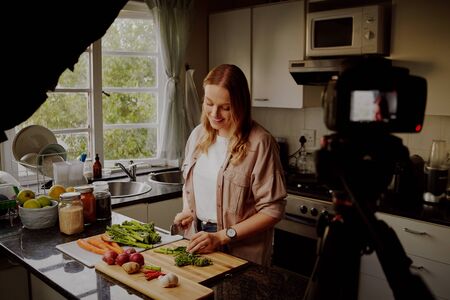 Woman in modern kitchen chopping fresh green vegetables to record food recipe on vlog - young woman vlogging in isolationの写真素材