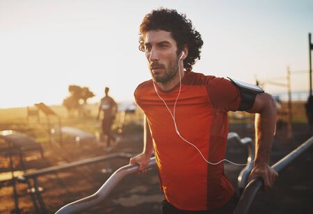 Portrait of an athletic young man listening to music on earphones doing exercise on parallel bars in the park - young man doing dips outdoorsの写真素材