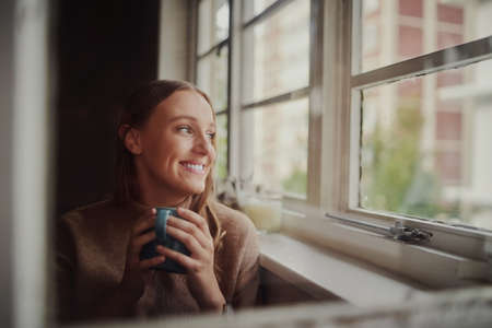 Quarantined young woman smiling drinking coffee and daydreaming looking out at empty streetsの写真素材