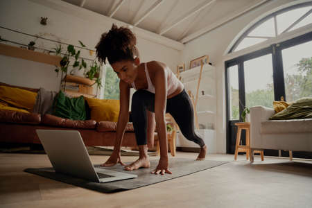 Happy woman watching video on laptop while exercising on yoga mat at home - lock down workoutの写真素材