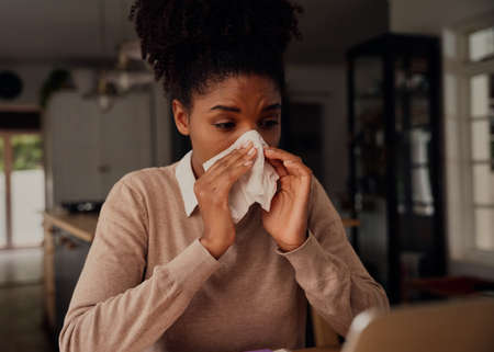 Sick woman blowing nose in tissue paper while sitting at home using laptop