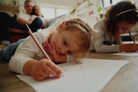 Portrait of two daughter lying on floor drawing artwork on paper at home with parents sitting on couch behind them - family bonding timeの写真素材