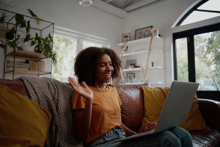 Smiling young african american woman relaxing on the couch waving hand while talking to friends on a...