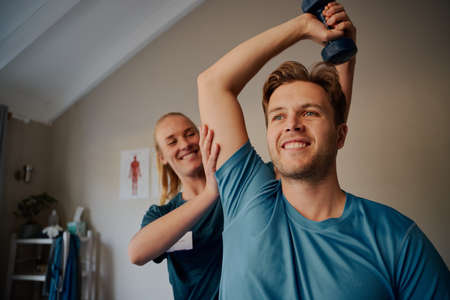 Portrait of handsome young smiling man exercising using dumbbells with female physiotherapistの写真素材