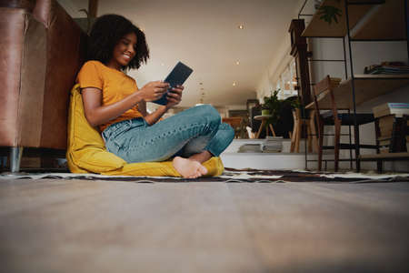 Low angle view of happy African American woman with afro sitting on floor with a pillow using a digital tabletの写真素材
