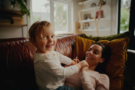 Portrait of excited little child playing with mother lying on couch at home during covid-19 lockdown