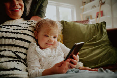 Smiling toddler child sitting with mother having fun using smartphone relaxing on couch