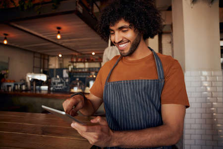Happy young man waiter wearing apron holding and using digital tablet in cafeの写真素材