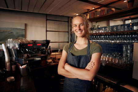Portrait of young female waitress wearing apron standing behind counter in cafe with crossed hands