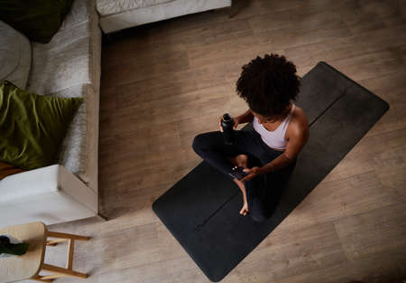 High angle view of young african woman holding bottle and using phone sitting on yoga matの写真素材