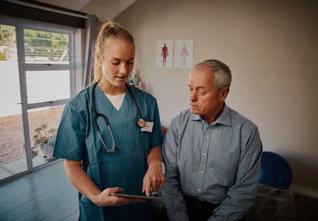 Portrait of young doctor in uniform and stethoscope holding digital tablet showing report to male...