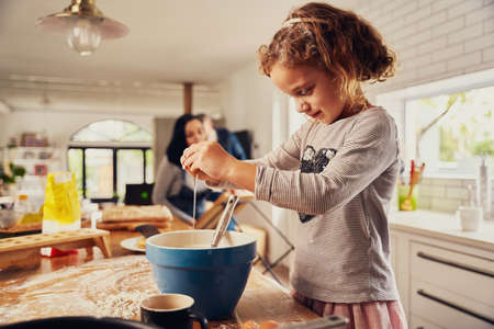 Cute little girl child breaking egg in a bowl while preparing dough in messy kitchen counter at homeの写真素材
