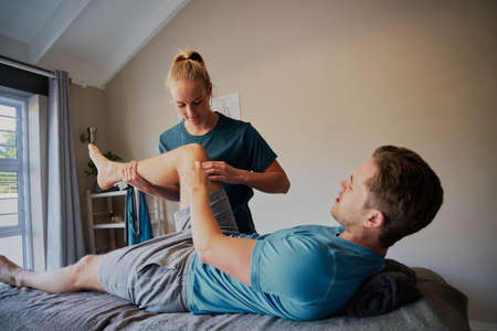 Young man lying on hospital bed showing joint pain position to young woman physiotherapistの写真素材