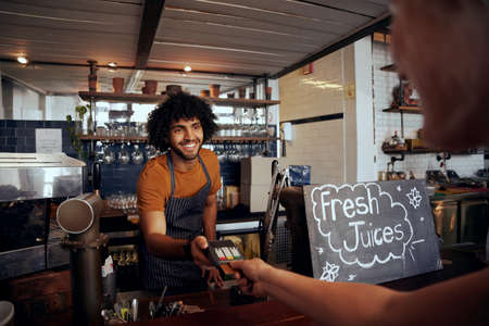 Portrait of smiling waiter holding credit card swipe machine while customer typing code in modern cafeの写真素材