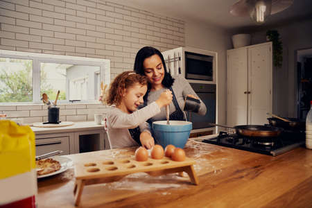 Smiling mother helping daughter mixing eggs and pouring water for dough in kitchen
