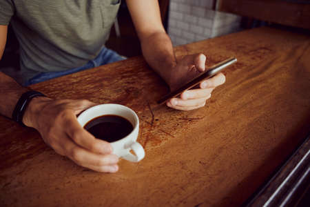 Closeup of male hands holding phone and coffee mug while sitting and relaxing in cafeの写真素材
