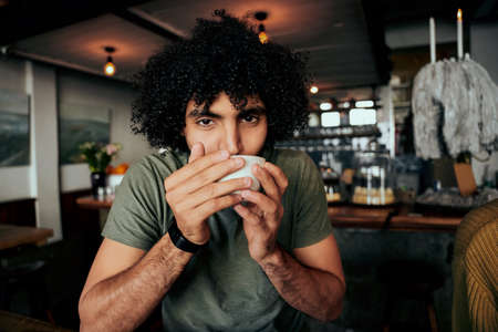 Handsome young man with curly hair sipping hot coffee relaxing in cafeの写真素材