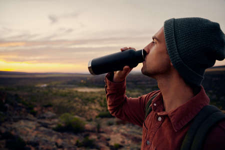 Tired male hiker wearing cap drinking beverage from bottle during early morning travel