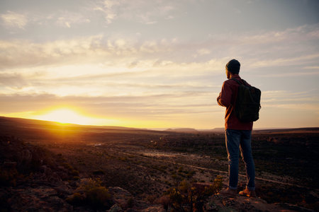 Low angle view of young male hiker with backpack standing on mountain top while hiking looking at...