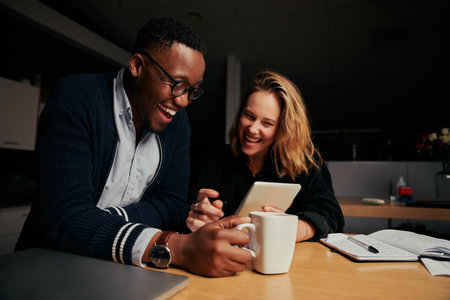 Happy businessman and businesswoman sitting together laughing while looking at digital tablet during break time at night - two colleagues smiling and working late togetherの写真素材
