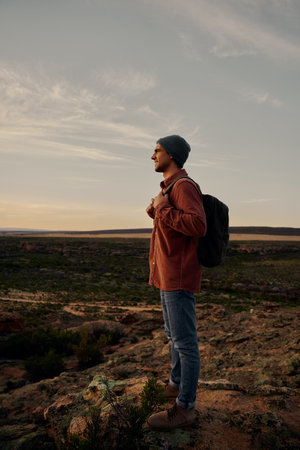 Successful hiker with bag and cap standing on mountain cliff looking at morning sunrise
