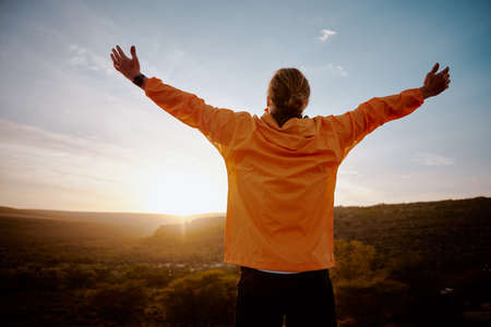 Rear view of young man standing on mountain with outstretched arms feeling relaxed after morning running in natureの写真素材