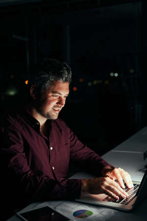Professional smiling focused businessman working with laptop computer late at night in dark officeの写真素材