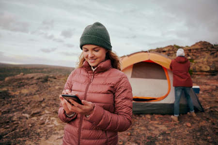 Happy young woman in winter clothing using smartphone during camping in mountain hill while man...