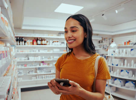 Young woman smiling while using smartphone to read prescription and chose medicine from shelf in pharmacyの写真素材