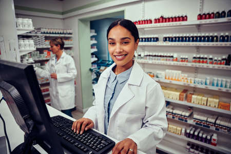 Portrait of smiling young female pharmacist wearing labcoat working on computer in drugstore looking at cameraの写真素材